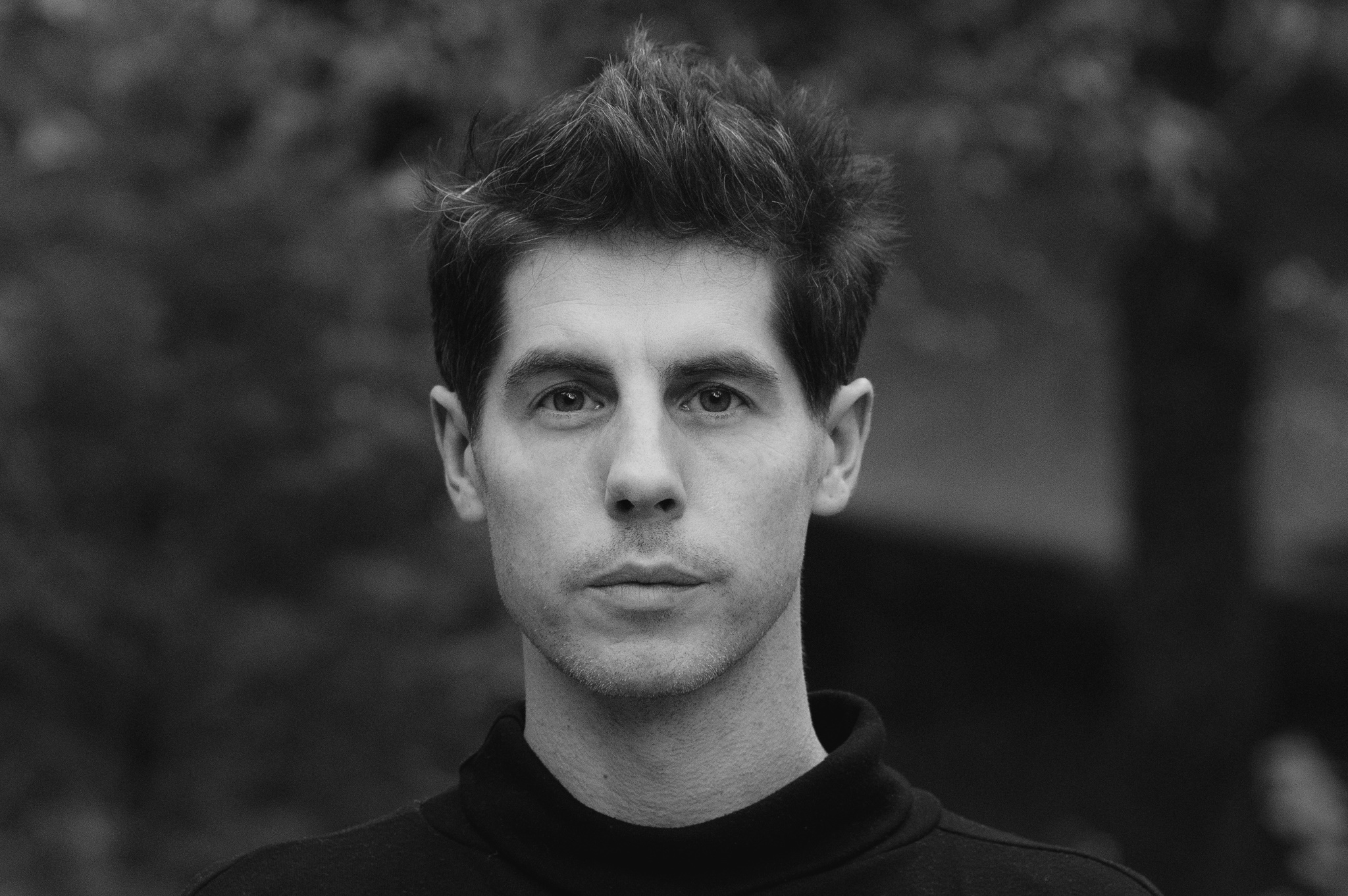 A black and white photo of the author in a black sweater, head and shoulders crop, against a wooded background